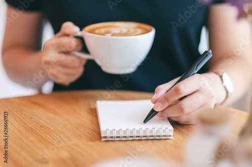 Closeup image of a woman writing on blank notebook while drinking coffee