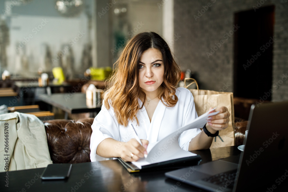 A young, sympathetic woman, not a thin-headed body building, sits in a cozy cafe, works at a computer, reviews documents. Business clothing style.