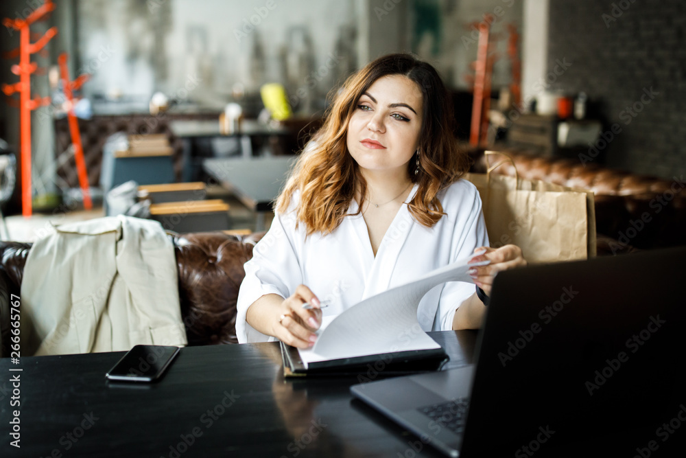 A young, sympathetic woman, not a thin-headed body building, sits in a cozy cafe, works at a computer, reviews documents. Business clothing style.
