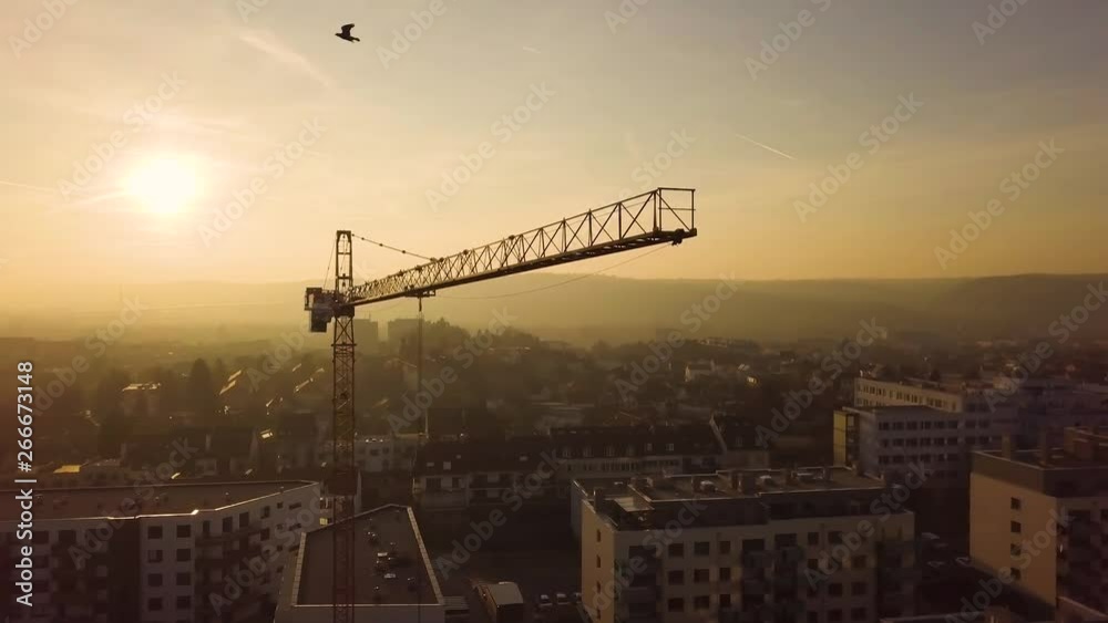 Aerial drone view of construction site crane over city at sunset with mountains in the background.