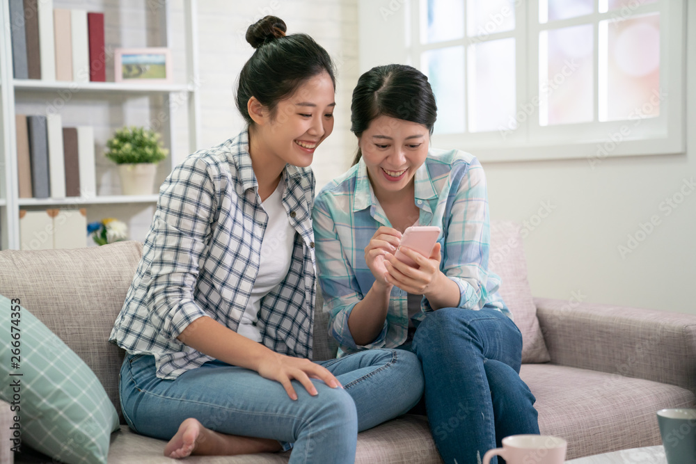 two young asian beautiful girls sitting on sofa in living room at home. women friends using smartphone watching recreational program funny videos online. korean female in couch shopping on internet