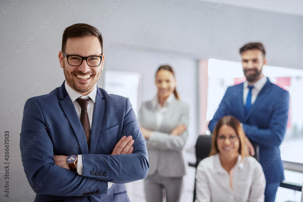Smiling successful Caucasian businessman in formal wear and with crossed arms standing in office. In background his team posing. Successful entrepreneurs are givers and not takers of positive energy. Smiling successful Caucasian businessman in formal wear and with crossed arms standing in office. In background his team posing. Successful entrepreneurs are givers and not takers of positive energy.
