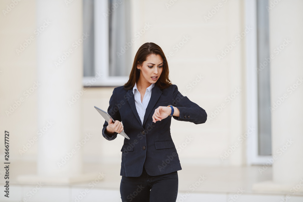 Young serious gorgeous brunette dressed smart casual looking at watch and holding tablet. Young serious gorgeous brunette dressed smart casual looking at watch and holding tablet.
