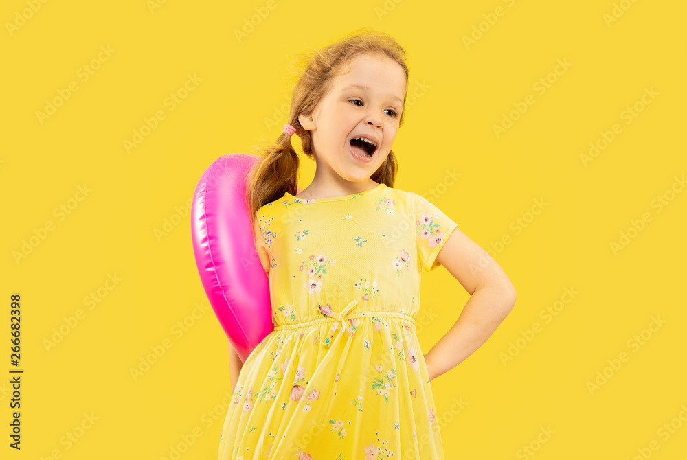 Beautiful emotional little girl isolated on yellow background. Half-lenght portrait of happy child wearing a dress and holding rubber pink flamingo. Concept of summer, human emotions, childhood. Beautiful emotional little girl isolated on yellow background. Half-lenght portrait of happy child wearing a dress and holding rubber pink flamingo. Concept of summer, human emotions, childhood.