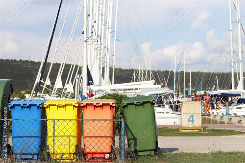 Multi-colored plastic garbage cans for waste sorting are on the quay in the marina against the backdrop of sailing yachts. Cleaning the yacht after the charter. Recycling and garbage disposal