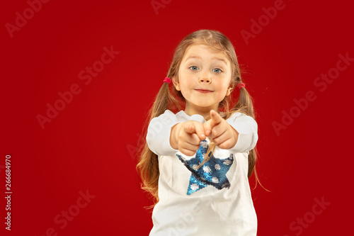 Beautiful emotional little girl isolated on red background. Half-lenght portrait of happy child showing a gesture and pointing up. Concept of facial expression  human emotions  childhood.