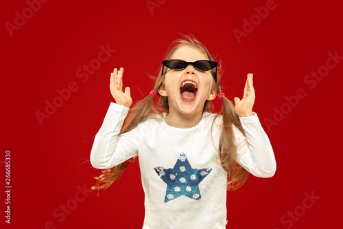 Beautiful emotional little girl isolated on red background. Half-lenght portrait of happy child showing a gesture and screaming. Concept of facial expression, human emotions, childhood.