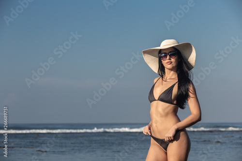 Beach vacation. Hot beautiful woman in sunhat and bikini standing with her arms raised to her head enjoying looking view of beach ocean on hot summer day.