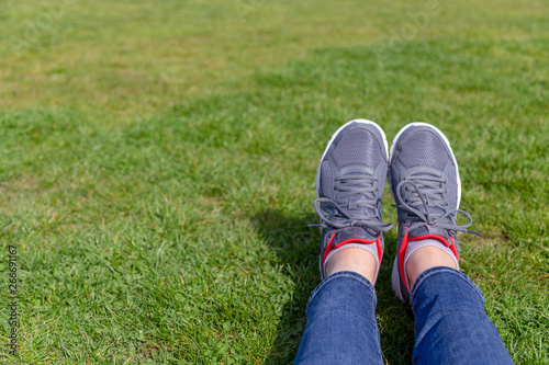 persons legs in jeans and grey trainer shoes laying on grass bright green grass with large copy space. Grey sneakers with bright trim, legs laying on grass in a relaxed manner.