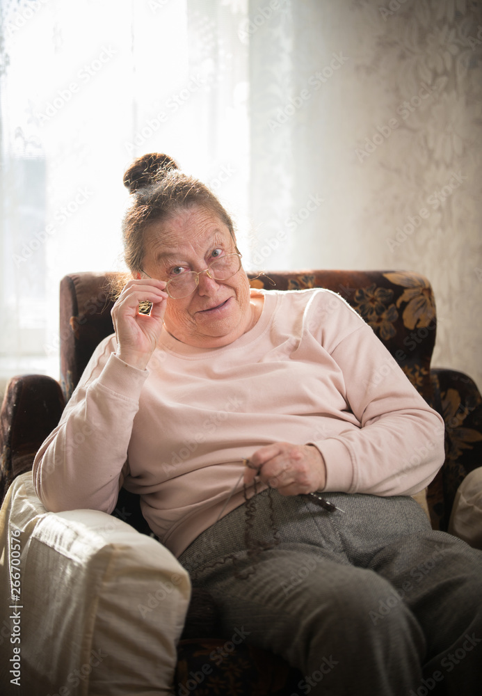 An old woman in glasses sitting in the armchair and put the glasses on the nose and looking at something An old woman in glasses sitting in the armchair and put the glasses on the nose and looking at something