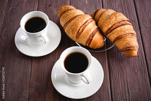 Two croissants on a saucer and two black coffees on a brown wooden table. 