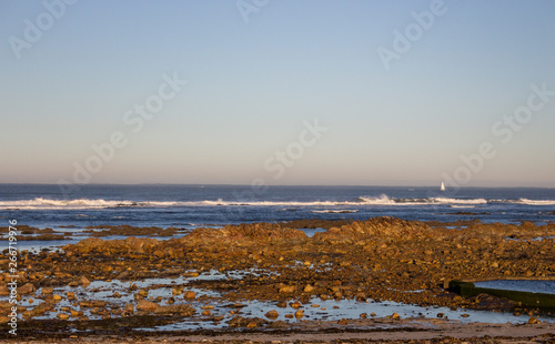 Rocky ocean coast with white sailing ship on horizon in the morning. Beautiful morning seascape. Stones and rocks on beach while outflow. Atlantic ocean landscape. Travel and tourism concept.