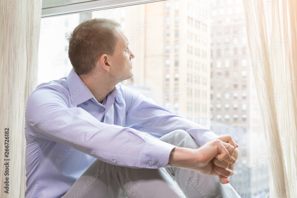 Young businessman man sitting dreaming on windowsill in dress shirt looking at New York City in Manhattan in hotel