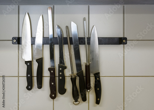 large kitchen knives and honing steels hanging on a magnetic holder on the tiled wall, copy space