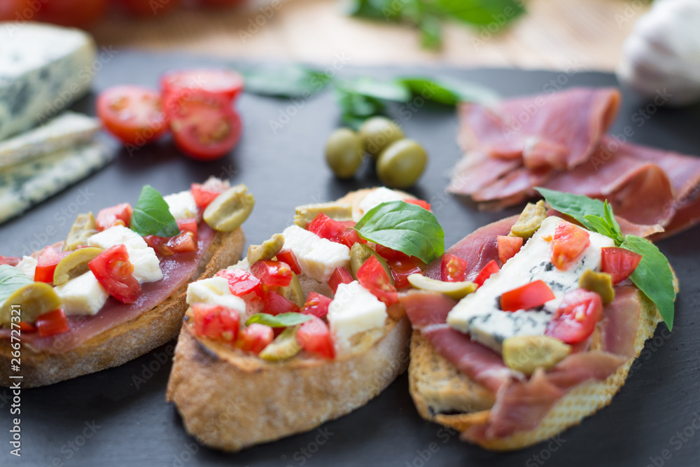 Traditional Italian bruschetta with blue cheese, feta, tomatoes, basil leaves, jamon on black stone background.