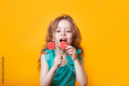 Funny child with candy lollipop  happy little girl eating big sugar lollipop on yellow bright background 