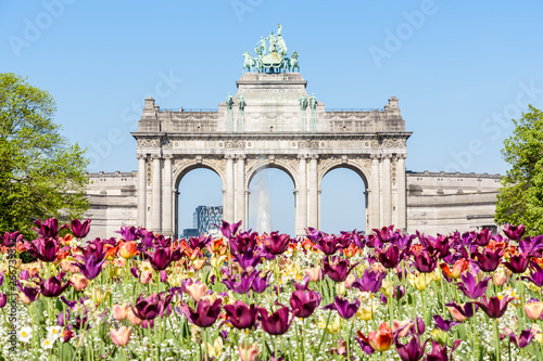 The arcade du Cinquantenaire, the triumphal arch erected by king Leopold II in the Cinquantenaire park in Brussels, Belgium, with flowers in bloom in the foreground against blue sky.