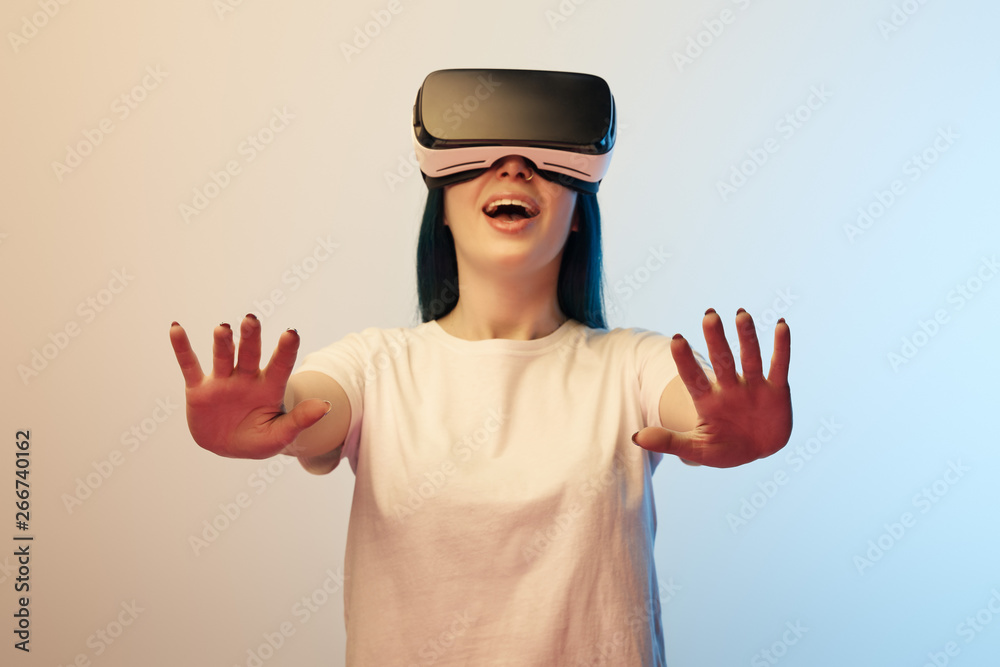 selective focus of cheerful woman in virtual reality headset gesturing on beige and blue selective focus of cheerful woman in virtual reality headset gesturing on beige and blue