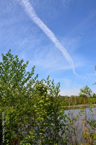 Kondesstreifen am blauen Himmel mit Wolken über Naturlandschaft am Wasser photo