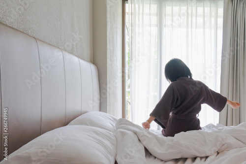 Woman stretching in bed after waking up, back view. Woman sitting near the big white window while stretching on bed after waking up with sunrise at morning, back view. © anon