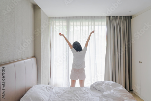 Fototapeta Naklejka Na Ścianę i Meble -  Woman stretching in bed after waking up, back view. Woman sitting near the big white window while stretching on bed after waking up with sunrise at morning, back view.