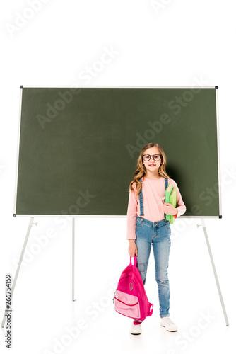 full length view of schoolgirl in glasses holding backpack and books near blackboard isolated on white
