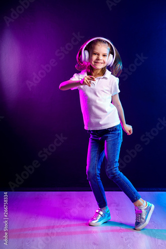 Girl of 7 years old listening to music in headphones and jump on dark colorful background . Dancing girl. Happy small girl dancing to music. Cute child enjoying happy dance music. © zamuruev