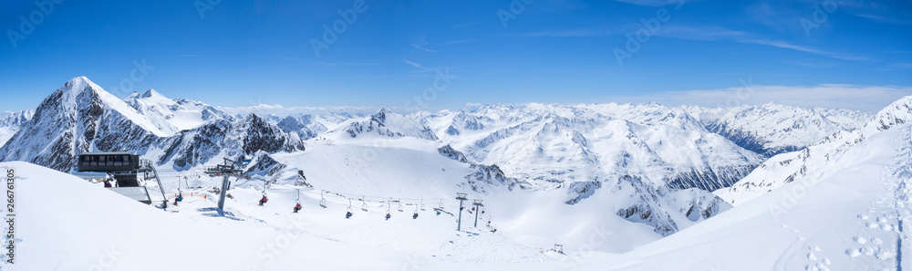 Panoramic landscape view from top of Wildspitz on winter landscape with snow covered mountain slopes and pistes and skiers on chair lift at Stubai Gletscher ski resort at spring sunny day. Blue sky