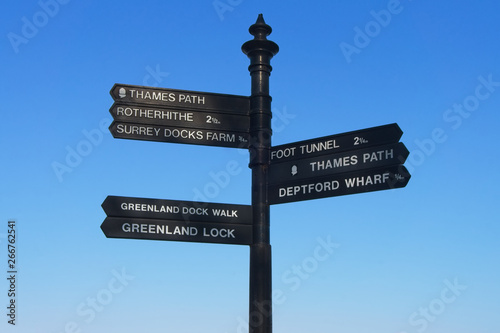 Black iron signpost giving directions to the Thames Path, Rotherhithe, Surrey Docks Farm, Greenland Dock Walk, Greenland Lock, Foot Tunnel, Thames Path, and Deptford Wharf in East London.