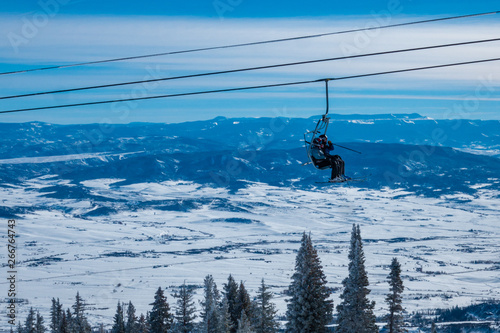 Skiers ascend the chairlift to the top of the ski slopes of Steamboat Springs, in the Rocky Mountains of Colorado, lined by Pine and Aspen trees. 