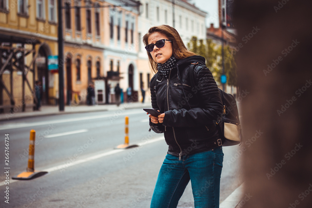 Brunette young woman wearing jeans, leather jacket and sunglasses – Girl with backpack hailing for a car or using phone to find directions and guidance during travel – Concept image for car sharing