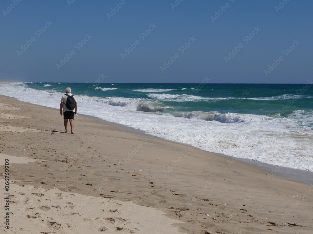 Older man with black backpack goes barefoot along the long, lonely sandy beach
