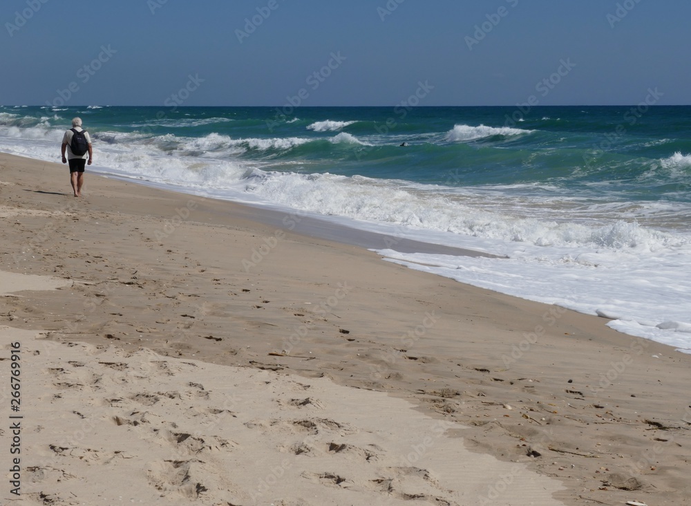 Older man with black backpack goes barefoot along the long, lonely sandy beach