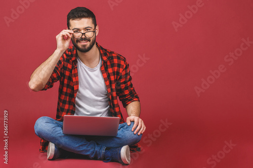 Should I write this post in my blog or not? Studio portrait of emotive handsome man with eyeglasses sitting with crossed legs on floor holding notebook on laps, isolated over red. © denis_vermenko