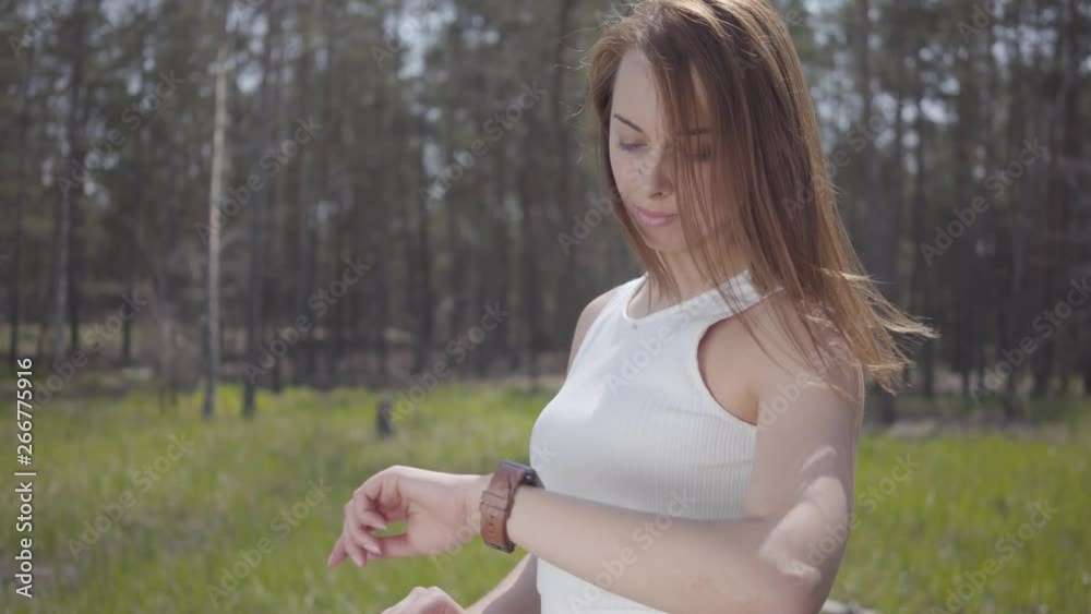 Beautiful woman with brown long hair looking at watch, waiting somebody on the green clearing around amazing forest Beautiful woman with brown long hair looking at watch, waiting somebody on the green clearing around amazing forest