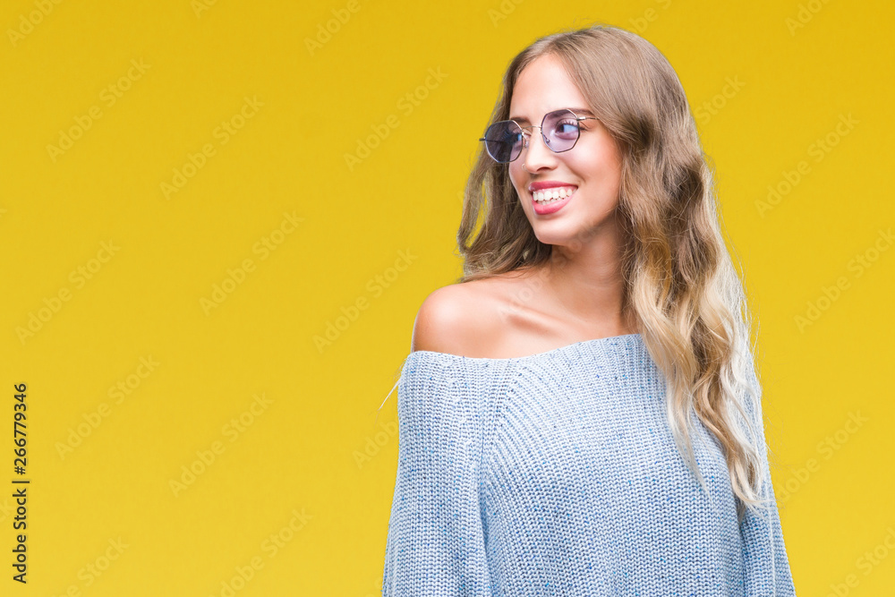 Beautiful young blonde woman wearing sunglasses over isolated background looking away to side with smile on face, natural expression. Laughing confident.