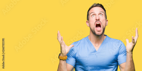 Handsome doctor man wearing medical uniform over isolated background crazy and mad shouting and yelling with aggressive expression and arms raised. Frustration concept.