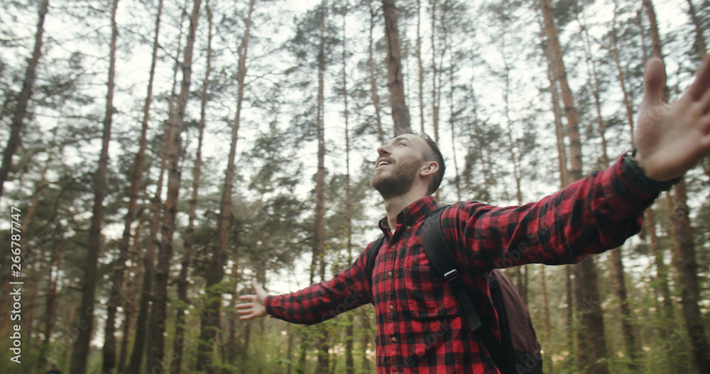Satisfied young bearded backpacked man dressed in checked shirt breathing healthy in the forest with raised up hands