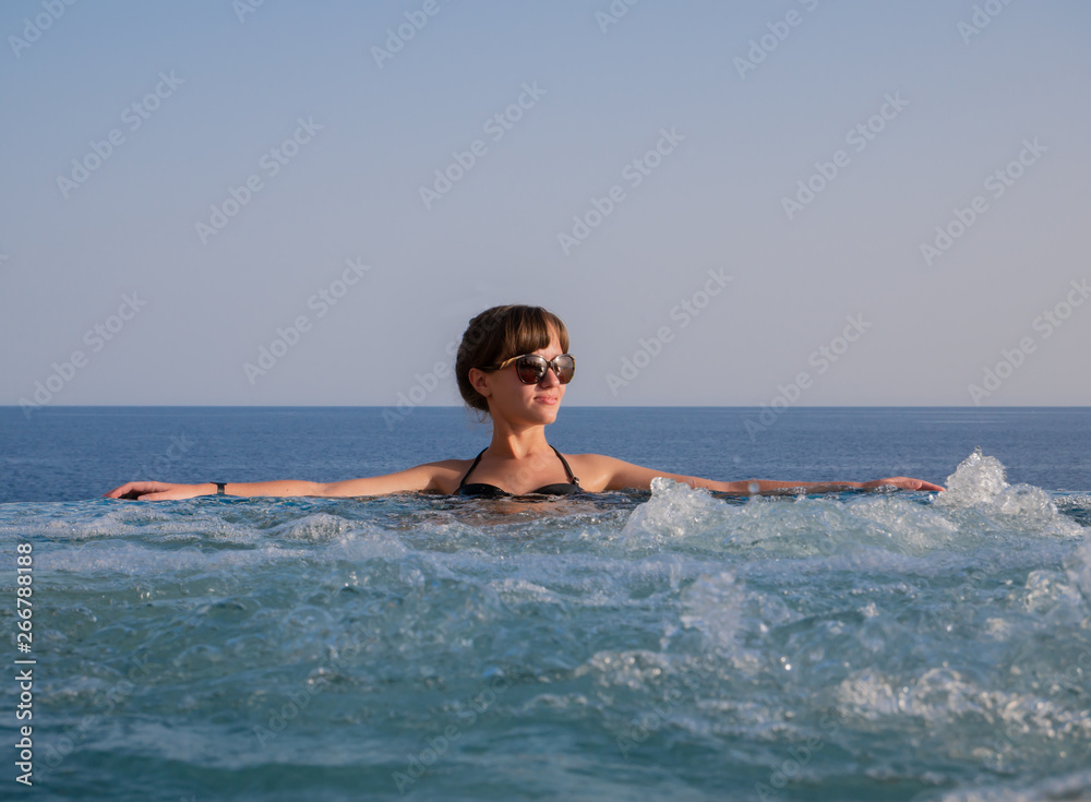 Pretty girl in sunglasses relaxing in the whirlpool pool at resort. Young brunette woman have relax in infinity jacuzzi against the background of the sea and sky.