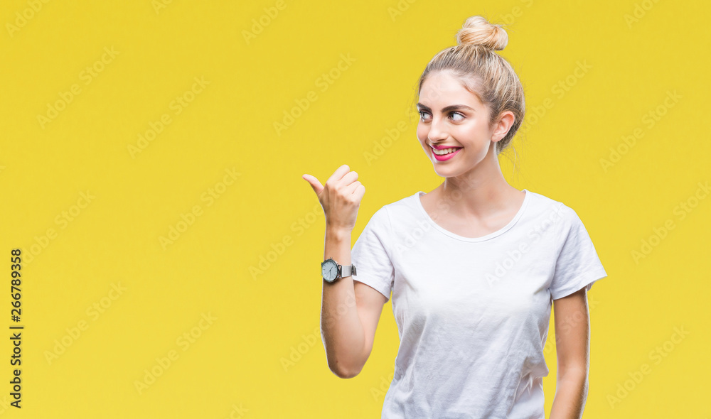 Young beautiful blonde woman wearing white t-shirt over isolated background smiling with happy face looking and pointing to the side with thumb up. Young beautiful blonde woman wearing white t-shirt over isolated background smiling with happy face looking and pointing to the side with thumb up.