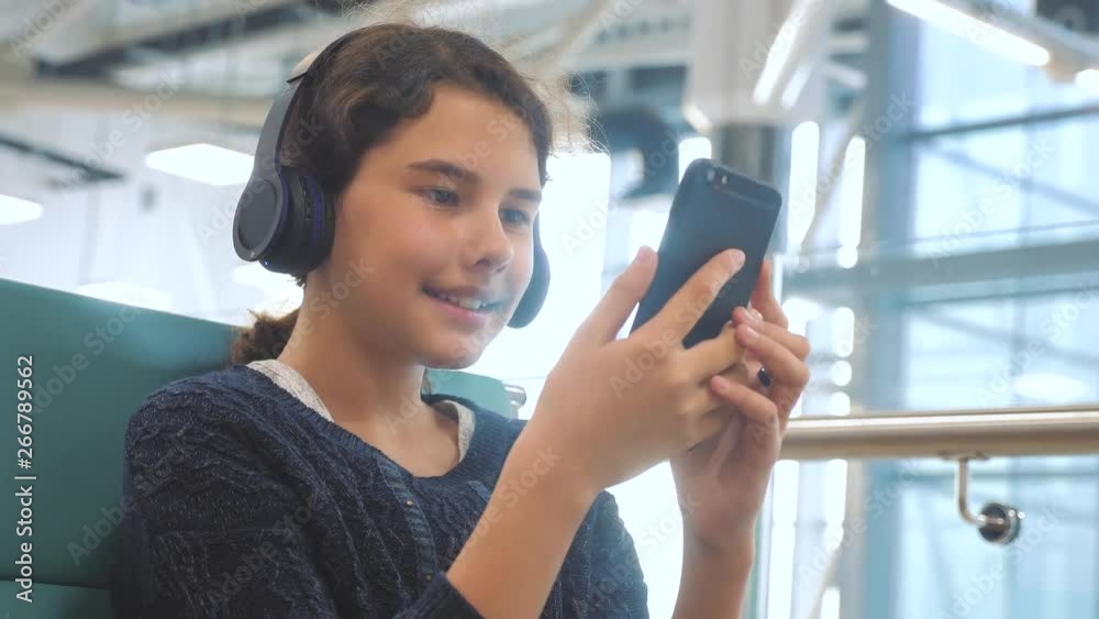 airport waiting hall room for a flight by plane. young happy teen girl in headphones listening to the music on smartphone chatting communicates in the messenger. teenager lifestyle girl in social airport waiting hall room for a flight by plane. young happy teen girl in headphones listening to the music on smartphone chatting communicates in the messenger. teenager lifestyle girl in social