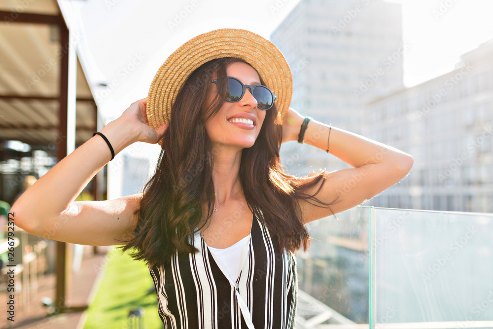 Close-up portrait of lovable caucasian woman in glasses touching her hat on city background in sunlight.