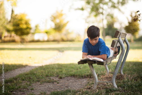 Giovane ragazzino sudamericano legge un libro sdraiato su una panchina nel parco sotto casa photo