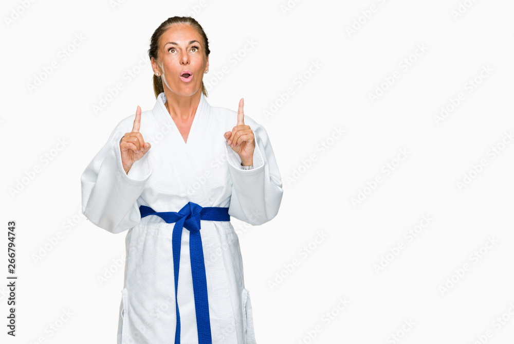 Middle age adult woman wearing karate kimono uniform over isolated background amazed and surprised looking up and pointing with fingers and raised arms. Middle age adult woman wearing karate kimono uniform over isolated background amazed and surprised looking up and pointing with fingers and raised arms.