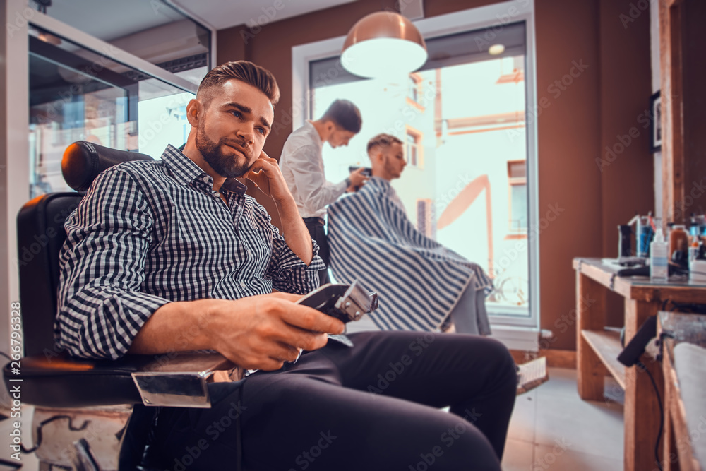 Young styilish man in checkered shirt is sitting at busy barbershop and holding trimmer for haircut. Young styilish man in checkered shirt is sitting at busy barbershop and holding trimmer for haircut.