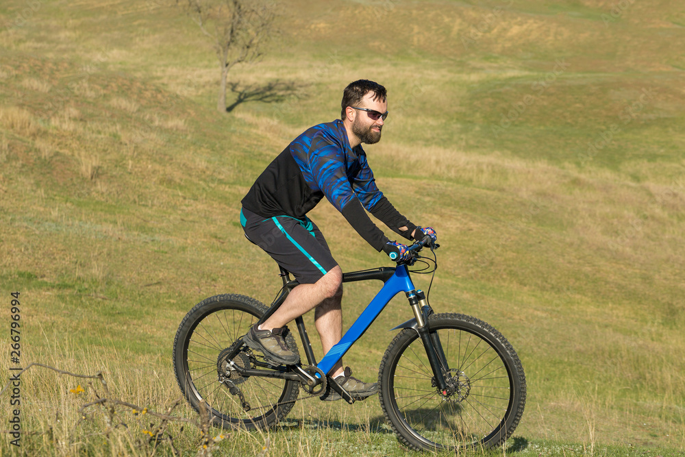 Cyclist in shorts and jersey on a modern carbon hardtail bike with an air suspension fork rides off-road on green hills near the forest Cyclist in shorts and jersey on a modern carbon hardtail bike with an air suspension fork rides off-road on green hills near the forest
