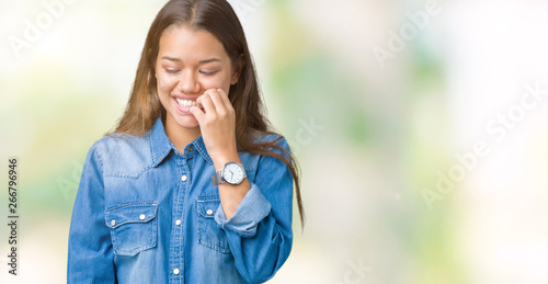 Young beautiful brunette woman wearing blue denim shirt over isolated background looking stressed and nervous with hands on mouth biting nails. Anxiety problem.