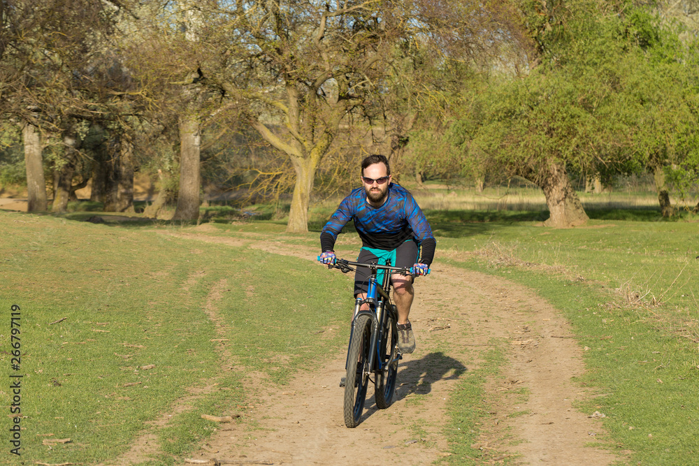 Cyclist in shorts and jersey on a modern carbon hardtail bike with an air suspension fork rides off-road on green hills near the forest	