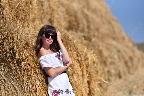 A young brunette girl in a white dress and glasses, against a background of a large haystack from round bales.