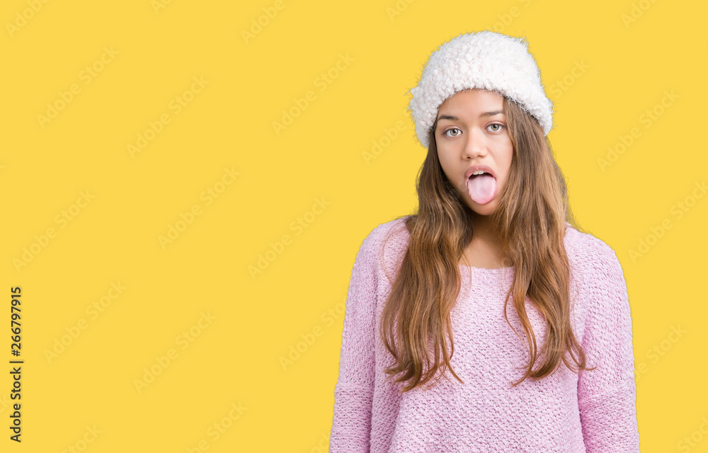 Young beautiful brunette woman wearing sweater and winter hat over isolated background sticking tongue out happy with funny expression. Emotion concept. Young beautiful brunette woman wearing sweater and winter hat over isolated background sticking tongue out happy with funny expression. Emotion concept.
